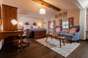 Ein gemütliches Schlafzimmer, das an einen ruhigen Rückzugsort erinnert, verfügt über ein Doppelbett mit gestreifter Bettwäsche, zwei gepolsterte Sessel und einen hölzernen Couchtisch. Das Zimmer ist perfekt für Flitterwöchner, die von ihrem Hochzeitstag träumen. Es verfügt über Hartholzböden, freiliegende Deckenbalken und gerahmte Bilder an der Wand.