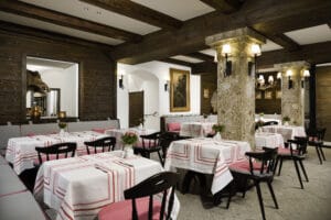 Ein gemütliches Restaurant-Interieur, perfekt für eine Hochzeit, mit Holzbalken und Steinsäulen. Die Tische sind mit weißen Tischdecken und roten Akzenten geschmückt. Schwarze Stühle mit rosa Kissen umgeben die Tische, während sanfte Beleuchtung eine warme, romantische Atmosphäre schafft, ideal für Hochzeiten.