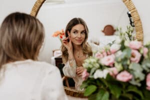 Eine Frau mit langen Haaren rückt ihre Ohrringe zurecht, bereitet sich auf ihre Hochzeit vor und schaut dabei in einen Spiegel. Im Vordergrund sind rosa und weiße Blumen zu sehen, die sich im Spiegel spiegeln und dem gemütlichen, sanft beleuchteten Raum eine romantische Note verleihen.