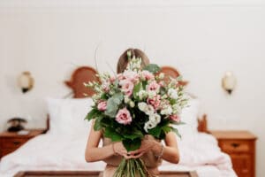In einem wunderschön dekorierten Schlafzimmer verbirgt eine Person ihr Gesicht hinter einem extravaganten Strauß rosa und weißer Blumen, perfekt für jede Hochzeit. Die üppigen grünen Blätter ergänzen die eleganten Holzmöbel und deuten auf eine sorgfältige Hochzeitsplanung hin.