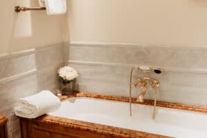 Ein gemütliches Badezimmer, perfekt zum Entspannen vor der Hochzeit, verfügt über eine weiße Badewanne mit Holzrahmen, einen eleganten goldenen Wasserhahn und ein ordentlich gefaltetes weißes Handtuch. Eine Vase mit weißen Rosen steht auf dem Wannenrand vor beige und weiß gefliesten Wänden.