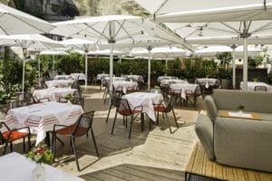 Die Außenterrasse des Restaurants ist ideal für eine Hochzeit und bietet hübsch arrangierte Tische, die mit weißen Tischdecken und kleinen Blumendekorationen geschmückt sind. Im Schatten großer weißer Sonnenschirme und umgeben von Grün ist sie mit ihren bequemen Sitzgelegenheiten perfekt für die Hochzeitsplanung.