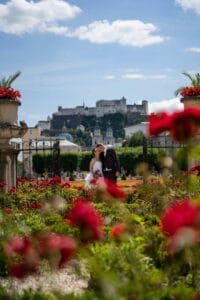 Ein Paar in Hochzeitskleidung steht inmitten leuchtend roter Blumen in einem Garten und feiert seine Hochzeit. Im Hintergrund krönt eine historische Festung den Hügel unter einem blauen Himmel mit vereinzelten Wolken.