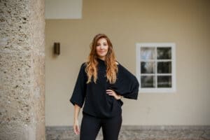 Eine Frau mit langen Haaren steht vor einer beigen Wand, die Hand auf der Hüfte, und stellt sich ihre perfekte Hochzeit vor. Sie trägt ein schwarzes Oberteil und eine schwarze Hose. Ein Fenster und eine Steinsäule im Hintergrund deuten auf eine elegante Hochzeitsplanung hin.