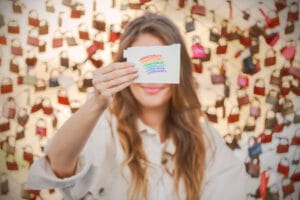 Eine Person hält ein Stück Papier mit einem handgezeichneten Regenbogen vor ihr Gesicht. Der Hintergrund erinnert an Heiratstraditionen und ist mit roten und braunen Locken gefüllt, die an einem Zaun hängen und Liebe und Hingabe symbolisieren, ähnlich einer sorgfältigen Hochzeitsplanung.