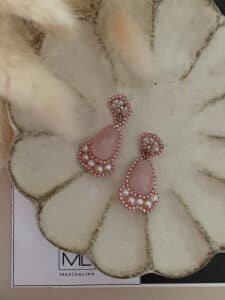 Ein Paar elegante Ohrringe mit rosafarbenen Steinen und Perlenakzenten, perfekt für eine Hochzeit, präsentiert auf einer dekorativen, gewellten Schale. Die Schale hat eine strukturierte, cremefarbene Oberfläche mit einem subtilen Glanz. Im Hintergrund sind weiche, verschwommene cremefarbene Blumen zu sehen.