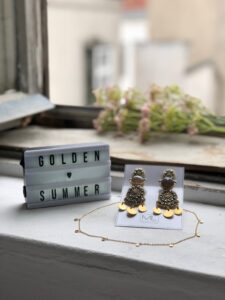 Ein Leuchtkasten mit dem Text „GOLDEN SUMMER“ steht auf einer Fensterbank neben eleganten baumelnden Ohrringen und einer zarten goldenen Halskette, perfekt für eine Hochzeit. Im Hintergrund verleihen verschwommene Blumen dieser bezaubernden Szene eine romantische Note.