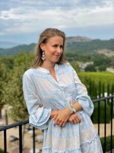 Eine Frau in einem blau-weißen Kleid, die vielleicht über ihre bevorstehende Hochzeit nachdenkt, steht an einem schwarzen Geländer mit malerischen Hügeln und Bäumen im Hintergrund. Sie trägt lange Ohrringe und eine Halskette, hat die Arme verschränkt und lächelt leicht, als würde sie von der Hochzeitsplanung träumen. Der Himmel ist teilweise bewölkt.