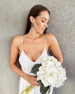 Eine Frau mit langen braunen Haaren trägt ein weißes Spitzenkleid, das an ein Hochzeitsensemble erinnert, und hält einen Strauß weißer Hortensien. Sie steht vor einer strukturierten grauen Wand und blickt nach rechts unten.