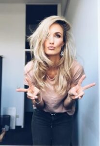 In einem hell erleuchteten Raum macht eine Person mit langen blonden Haaren, einem rosa Oberteil und dunklen Hosen ein verspieltes Schmollgesicht und hält dabei Friedenszeichen hoch. Es ist, als würde sie etwas Besonderes feiern – vielleicht ihre eigene Hochzeitsplanung.