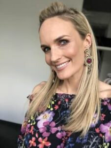 Eine lächelnde Frau mit langen blonden Haaren, geschmückt mit großen goldenen und roten Ohrringen und einem schulterfreien Blumentop, strahlt in einem Innenbereich vor einem unscharfen Hintergrund Freude aus und feiert vielleicht ihre Hochzeit.
