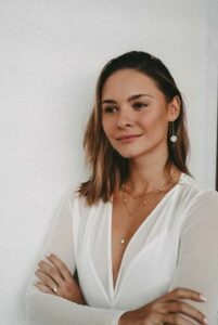 Eine Frau mit schulterlangem Haar steht vor einer weißen Wand. Sie trägt ein weißes Oberteil und Schmuck. Sie lächelt leicht und hat die Arme verschränkt. Vielleicht denkt sie über ihre bevorstehende Hochzeit nach oder über die Freuden der Hochzeitsplanung.