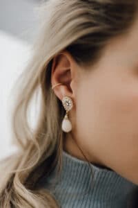 Nahaufnahme einer Frau, die einen Perlenohrring mit einem funkelnden runden Ohrstecker trägt, perfekt für eine elegante Hochzeit. Ihr blondes Haar ist hinter ihr Ohr gesteckt und sie trägt einen grauen Rollkragenpullover.