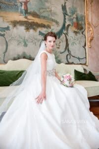 Eine Braut in einem weißen Brautkleid sitzt auf einem Vintage-Sofa und verkörpert die Essenz der Hochzeits-Eleganz. Sie hält einen Blumenstrauß in einem eleganten Raum, der mit einem dekorativen Wandgemälde geschmückt ist. Ihr Kleid hat ein tailliertes Mieder und einen weiten Rock, ergänzt durch einen langen Schleier.