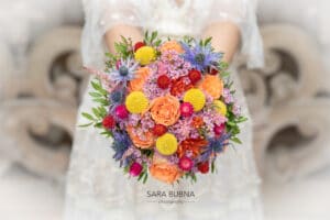 Eine Braut, vertieft in die Freuden der Hochzeitsplanung, hält einen bunten Strauß mit orangefarbenen Rosen und leuchtend gelben, roten und violetten Blumen, ergänzt durch üppiges grünes Laub. Der Strauß steht im Mittelpunkt des Bildes, mit verschwommenen, kunstvollen Hintergrunddetails, die diesen Hochzeitsmoment noch verstärken.