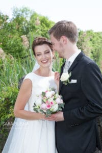 Braut und Bräutigam lächeln freudig an ihrem Hochzeitstag. Die Braut hält einen Strauß rosafarbener und weißer Blumen in der Hand und trägt ein weißes Kleid, während der Bräutigam einen schwarzen Anzug mit einer Ansteckblume aus weißen Rosen trägt. Sie sind draußen mit viel Grün im Hintergrund.