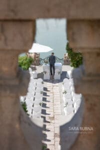 Ein Blick durch eine Steinbalustrade zeigt eine Hochzeitsdekoration im Freien mit weißen Stühlen, die in Reihen mit Blick auf ein Gewässer angeordnet sind. Eine Person steht neben einem dekorativen Geländer unter einem weißen Sonnenschirm. Die Szene ist friedlich und elegant arrangiert und spiegelt das Herz sorgfältiger Hochzeitsplanung wider.