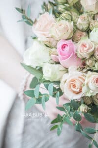 Ein Strauß pastellfarbener Rosen mit rosa, pfirsichfarbenen und weißen Blüten wird sanft in jemandes Hand gehalten. Durch das Grün noch schöner, strahlt das Arrangement eine sanfte romantische Atmosphäre aus – perfekt für eine Hochzeitsfeier.