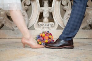 Ein Frauenfuß in einem rosa Absatzschuh und ein Männerfuß in einem braunen Schuh stehen Zehe an Zehe auf einem gefliesten Boden, perfekt für ihre Hochzeit. Zwischen ihnen liegt ein bunter Blumenstrauß mit roten, rosa, orangen und violetten Blumen. Im Hintergrund ist kunstvolles Mauerwerk zu sehen.