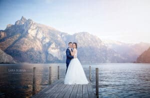 Braut und Bräutigam stehen auf einem Holzsteg und umarmen sich während ihrer Hochzeit. Hinter ihnen liegt ein ruhiger See, der von majestätischen Bergen unter einem klaren Himmel gesäumt ist. Die Umgebung ist friedlich und malerisch, ideal für diejenigen, die davon träumen, inmitten der Schönheit der Natur zu heiraten.