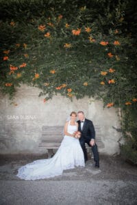 Braut und Bräutigam sitzen in glückseliger Ehe auf einer Holzbank, umgeben von Grün und leuchtend orangefarbenen Blumen. Die Braut in einem weißen Kleid mit Blumenstrauß und der Bräutigam in einem eleganten schwarzen Anzug lächeln strahlend in ihrer idyllischen Umgebung im Freien.