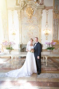 Ein Paar in Hochzeitskleidung steht zusammen in einem reich verzierten Raum mit dekorativem Hintergrund und verkörpert die Freude ihrer Hochzeit. Die Braut hält einen Blumenstrauß und beide lächeln. Sie sind von elegantem Dekor mit Blumenarrangements und aufwendigen Wanddesigns umgeben.