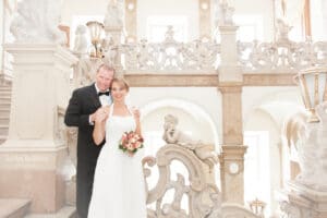 Braut und Bräutigam posieren auf einer verzierten Treppe in einer prachtvollen Kulisse und verkörpern damit die Eleganz, die beim Heiraten im Mittelpunkt steht. Die Braut, in einem weißen Kleid und mit einem Blumenstrauß in der Hand, steht neben ihrem Bräutigam im eleganten schwarzen Smoking. Marmorstatuen und kunstvolle Geländer unterstreichen die anspruchsvolle Kulisse dieser bedeutungsvollen Hochzeit.