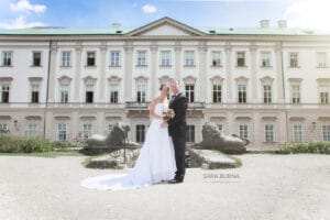 Braut und Bräutigam stehen gemeinsam vor einem großen, historischen Gebäude mit grünem Dach und feiern ihre Hochzeit. Sie lächeln und umarmen sich, während die Braut einen Blumenstrauß hält. Zwei Löwenstatuen flankieren sie im Vordergrund unter einem klaren, sonnigen Himmel.