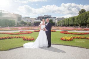 Ein Paar im Hochzeitskleid steht in einem Garten mit bunten Blumenbeeten und verkörpert eine perfekte Hochzeitsszene. Die Braut strahlt in ihrem weißen Kleid, während der Bräutigam in seinem schwarzen Smoking schick aussieht. Ein historisches Gebäude und ein teilweise bewölkter Himmel tragen zur bezaubernden Atmosphäre ihres besonderen Tages bei.