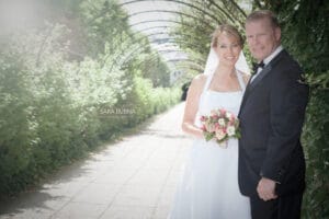 Braut und Bräutigam posieren unter einem grünen Torbogen und verkörpern die Freude des Heiratens. Die Braut hält einen Blumenstrauß und beide lächeln herzlich in die Kamera. Das Sonnenlicht fällt durch und schafft eine sanfte, romantische Atmosphäre, die perfekt für jede durchdachte Hochzeitsplanung ist.