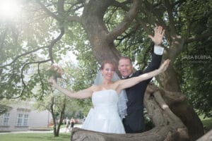 Ein lächelndes Paar im Hochzeitskleid posiert verspielt unter einem großen, gewundenen Baum und verkörpert die Freude des „Heiraten“. Die Braut hält einen Blumenstrauß und beide haben ihre Arme ausgestreckt. Sonnenlicht fällt durch die Blätter und schafft eine verträumte Atmosphäre, perfekt für ihre Hochzeit.