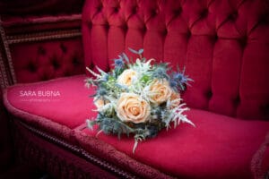 Ein Strauß pfirsichfarbener Rosen und Grünpflanzen ruht auf einem plüschigen, roten Sitzpolster, perfekt für eine elegante Hochzeit. Die leuchtenden Farben schaffen einen auffälligen Kontrast.