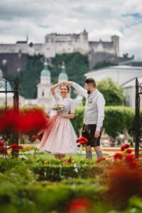 Ein Paar in traditioneller Kleidung steht in einem lebhaften Garten und feiert seine Hochzeit. Der Mann wirbelt die Frau unter einem Torbogen herum, im Hintergrund ist eine historische Festung zu sehen. Leuchtend rote Blumen im Vordergrund sorgen für eine lebendige und romantische Szene.