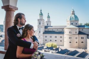 Ein Paar in formeller Kleidung steht auf einem Balkon, wobei der Mann die Frau von hinten umarmt. Sie blicken auf eine Stadtlandschaft mit Kuppelgebäuden unter einem klaren blauen Himmel. Die Frau, die einen Blumenstrauß hält, denkt über ihre Hochzeit nach und die Freude des Heiratens inmitten dieser Schönheit.