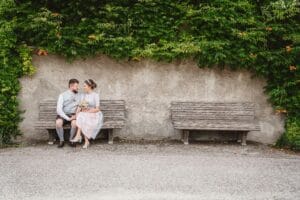 Ein Paar in traditioneller Kleidung sitzt auf einer Holzbank vor einer efeubewachsenen Wand, wobei die Frau einen Blumenstrauß hält – eine Szene, die an eine verträumte Hochzeit erinnert. Eine weitere leere Bank steht neben ihnen und verstärkt die friedliche und romantische Atmosphäre.