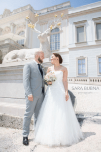 Die Braut im weißen Kleid und der Bräutigam im hellgrauen Anzug stehen lächelnd vor einer Hirschstatue mit Sternenkrone und fangen die Essenz ihres Hochzeitstages an diesem sonnigen Tag ein.
