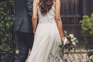 Braut und Bräutigam gehen gemeinsam an ihrem Hochzeitstag spazieren. Die Braut trägt ein weißes Kleid und hält einen Strauß weißer Blumen, während der Bräutigam einen dunklen Anzug trägt. Sie sind von üppigem Grün umgeben, im Vordergrund sind Lavendelblüten zu sehen.