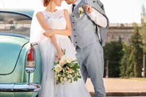 Braut und Bräutigam stehen neben einem blauen Oldtimer, wobei der Bräutigam verspielt sein Jackett hält. Die Braut hält einen Strauß weißer und cremefarbener Blumen in der Hand. Beide, in eleganter Hochzeitskleidung, zeigen ihre fröhliche Stimmung vor der verschwommenen Außenkulisse – ein perfekter Moment in ihrer Hochzeitsplanung.