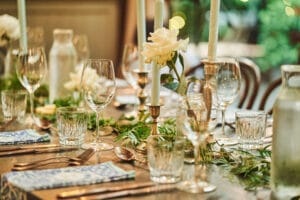 Ein wunderschön gedeckter Esstisch mit elegantem Glasgeschirr, Silberbesteck, blauen Servietten und großen weißen Kerzen, umgeben von Grünpflanzen und weißen Rosen. Die warme und einladende Atmosphäre lässt auf eine intime Hochzeitsfeier oder ein Fest schließen.