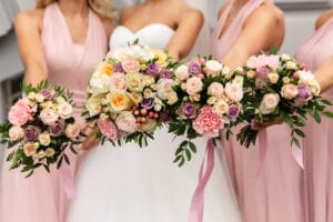 Vier Brautjungfern in rosa Kleidern halten Sträuße aus Rosen in Pfirsich-, Lila- und Weißtönen. In der Mitte steht eine Braut in ihrem eleganten weißen Kleid und hält ebenfalls einen ähnlichen Strauß, was der Hochzeit eine verträumte Note verleiht.