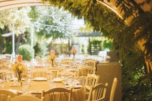 Ein bezaubernder Hochzeitsempfangsbereich im Freien bietet runde Tische mit weißen Tischdecken, umgeben von eleganten weißen Stühlen. Jeder Tisch ist mit einem farbenfrohen Blumenschmuck geschmückt, während sanftes, natürliches Licht durch das üppige Grün fällt – eine Traumkulisse für alle, die schon weit mit ihrer Hochzeitsplanung sind.