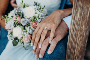 Eine Nahaufnahme der Hände eines Paares, die auf einer Holzoberfläche ruhen. Die Frau im Hochzeitskleid zeigt einen Diamantring, der ihre Entscheidung zu heiraten symbolisiert. Sie hält einen Strauß blassrosa, weißer und lavendelfarbener Rosen mit Grünpflanzen und Schleierkraut in der Hand und fängt damit die Essenz der Hochzeitsplanung ein.