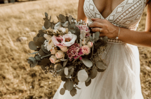 Auf einer sonnenbeschienenen Wiese hält eine Braut in einem ärmellosen Spitzenbrautkleid einen Strauß rosa und weißer Blumen mit Eukalyptusblättern in den Händen. Ihr Armband und ihr Ehering glitzern sanft, während das Licht die filigranen Details ihres Kleides hervorhebt – ein perfekter Moment in ihrer Hochzeitsplanung.