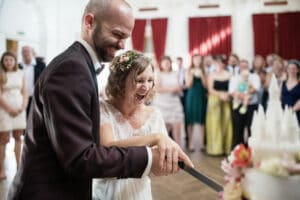 Braut und Bräutigam schneiden während ihrer Hochzeit freudig gemeinsam ihre Hochzeitstorte an. Die Braut trägt einen Blumenkopfschmuck und ein Spitzenkleid, während der Bräutigam einen dunklen Anzug trägt. Im Hintergrund stehen Gäste in Abendgarderobe, einige halten Kinder, in einem Raum mit roten Vorhängen.