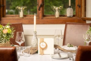 Ein für ein Essen gedeckter Tisch mit elegantem Dekor, einschließlich Weingläsern, einer Kerze, einem Blumenarrangement und einer Menükarte. Das Ambiente im warm beleuchteten Speisezimmer ist perfekt für eine Hochzeit und verfügt über einen Holzfensterrahmen, durch den man das Grün draußen sehen kann.