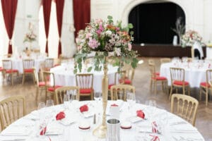 Ein geräumiger, eleganter Bankettsaal, perfekt für eine Hochzeit, mit runden Tischen, die für die Veranstaltung gedeckt sind. Jeder Tisch ist mit hohen Blumengestecken und roten und weißen Dekorationen geschmückt und von goldenen Stühlen mit roten Kissen umgeben. Der Hintergrund ist mit tiefroten Vorhängen versehen, ideal für Ihre Hochzeitsfeier.