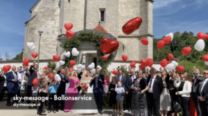Eine Hochzeitsfeier mit Gästen vor einem historischen Gebäude schafft eine bezaubernde Atmosphäre, während die Menschen rote und weiße herzförmige Luftballons in den Himmel steigen lassen. Der festliche Tag ist Ausdruck einer sorgfältigen Hochzeitsplanung, und alle sind elegant und in formeller Kleidung gekleidet.