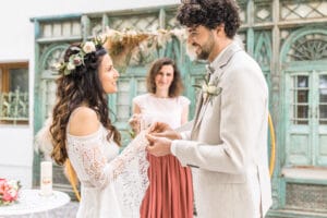 Ein Paar steht sich gegenüber und hält sich bei einer wunderschönen Hochzeitszeremonie an den Händen. Die Braut in einem Spitzenkleid und einem Blumenkopfschmuck ergänzt den hellen Anzug des Bräutigams. Eine Frau lächelt herzlich hinter ihnen, während sie vor verzierten grünen Türen stehen und ihren Hochzeitsmoment unvergesslich machen.