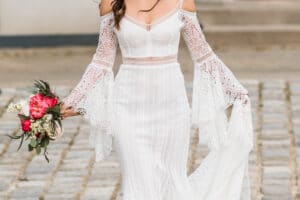 Eine Frau in einem fließenden weißen Spitzenbrautkleid mit schulterfreien Ärmeln, das die Essenz einer Hochzeit verkörpert, hält einen Strauß rosa und weißer Blumen, während sie anmutig über das Kopfsteinpflaster geht.