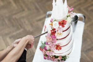 Ein Paar feiert seine Hochzeit und schneidet eine hohe, weiße Hochzeitstorte an, die mit rosa und roten Blumen geschmückt ist. Die Torte, anmutig auf einem mit einer weißen Tischdecke bedeckten Tablett platziert, spiegelt ihre sorgfältige Hochzeitsplanung wider. Hände, die das Messer halten, sind vor dem Hintergrund eines Holzbodens zu sehen.
